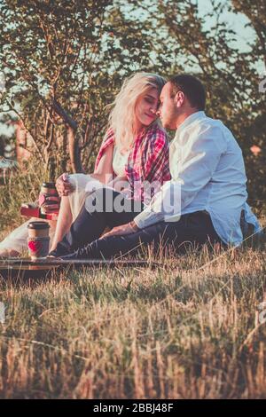 Couple romantique embrassant tout en pique-nique au coucher du soleil. L'homme en chemise blanche et la jeune femme séduisante sont assis ensemble sur l'herbe sur le Banque D'Images