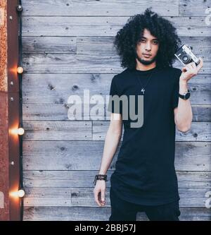 Homme africain américain avec des cheveux afro typiques posant avec un appareil photo vintage sur fond de vieux planches. Vêtu d'une tenue noire. Banque D'Images