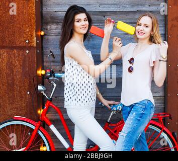 Deux petites amies habillées de tenues tendance, un Jean, des hauts se reposent, mangent de la glace assise sur un vélo rouge vintage tendance. Extérieur. Banque D'Images