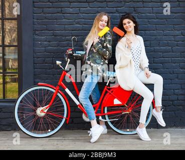 Deux amies déguisés en tenues tendance se reposent, mangent de la glace assise sur un vélo rouge vintage tendance Banque D'Images