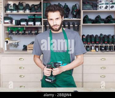 Un hipster barbu dans des vêtements de travail, un T-shirt et un tablier vert, tient un tournevis sans fil dans ses mains Banque D'Images