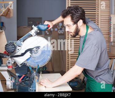 Jeune homme de barbe hipster dans des combinaisons noires par profession menuisier constructeur avec une scie circulaire un panneau en bois sur une table en bois dans l'atelier Banque D'Images