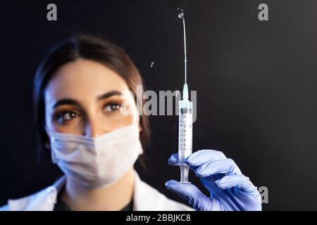 Blanc caucasien Doctor ou infirmière femme avec masque et gants d'examen bleus tient la seringue d'injection et le vaccin et l'écurent sur un fond noir Banque D'Images