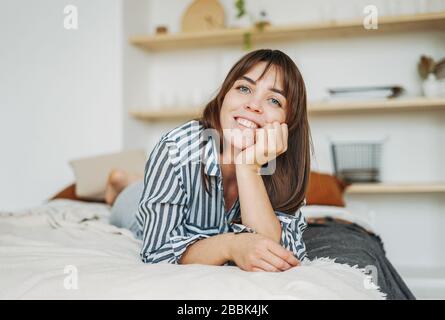 Jeune femme heureuse brunette dans des vêtements décontractés reposer sur le lit à l'intérieur lumineux Banque D'Images