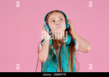 Une petite fille avec casque explose la gomme à mâcher rose sur un fond rose. Banque D'Images