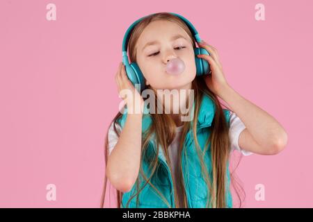 Une petite fille avec casque explose la gomme à mâcher rose sur un fond rose. Banque D'Images