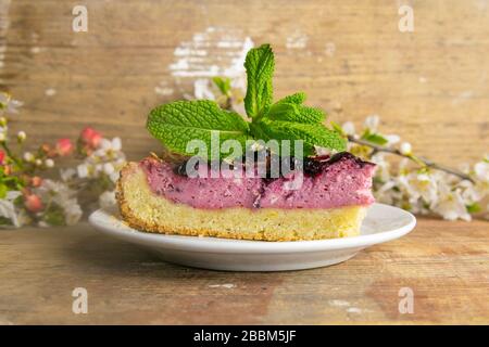 Des tranches de mousse de myrtilles avec des baies décorées de verglas miroir et des feuilles de menthe sur une plaque blanche avec des fleurs blanches sont sur table en bois, à proximité Banque D'Images