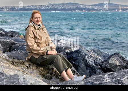 Jeune femme caucasienne souriante, vêtue de vêtements d'hiver chauds, se trouve sur la rive de la baie de la Corne d'Or, en toile de fond des vagues de tempête et de la c Banque D'Images