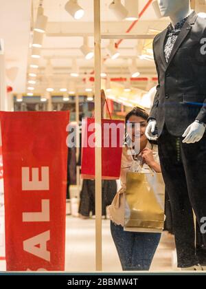 Belle femme dans des vêtements à la mode montre des sacs de shopping sur des vitrines de magasin.signe rouge sur une fenêtre de magasin. Vente dans un magasin de vêtements. Banque D'Images