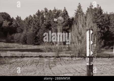 Sépia tonation de clôture de fil barbelé sur un poteau T en premier plan un cadre rural d'automne avec moulin à vent en arrière-plan Banque D'Images