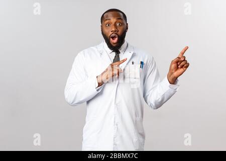 J'ai impressionné le médecin afro-américain masculin de bonne qualité en manteau blanc, pointant vers le coin supérieur droit, éveiller et affamer la caméra excitée et émerveillée, dire Banque D'Images