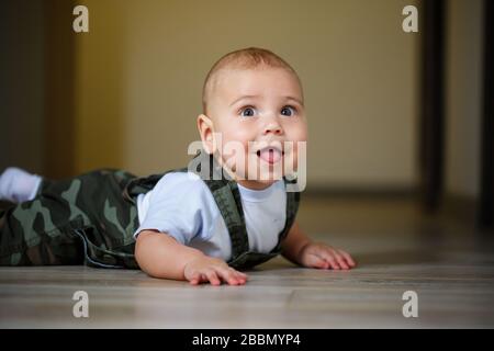 petit garçon de 8 mois en salopette, chemise blanche et chaussettes blanches rampant sur le sol et souriant