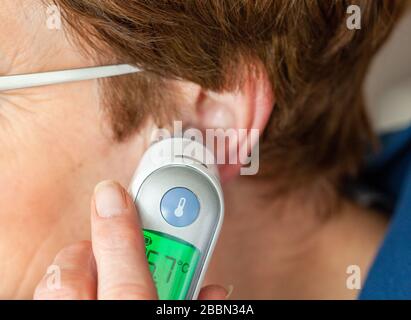 Femme prenant sa propre température avec un thermomètre électronique dans l'oreille. Peut être utilisé pour indiquer des concepts allant de l'auto-diagnostic pour des maladies telles que le Coronavirus, le COVID-19 à l'auto-isolation.ou l'autonomie Banque D'Images