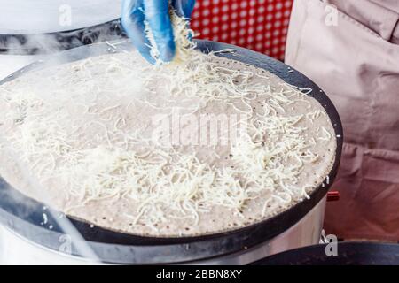 Faire des crêpes crêpes à un marché alimentaire. Une main fait des crêpes salées à l'extérieur ajoutant du fromage râpé à la crêpe sur une grille métallique. Banque D'Images
