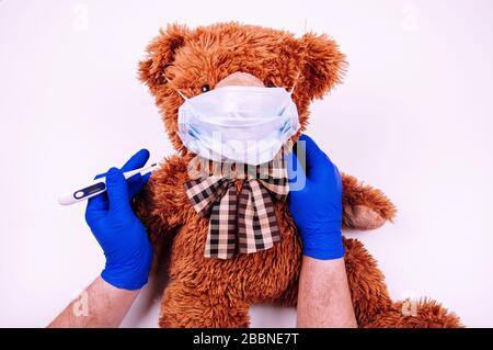 Les mains de l'homme dans des gants bleus d'élimination contiennent du thermometr et du masque médical sur l'ours en peluche Banque D'Images