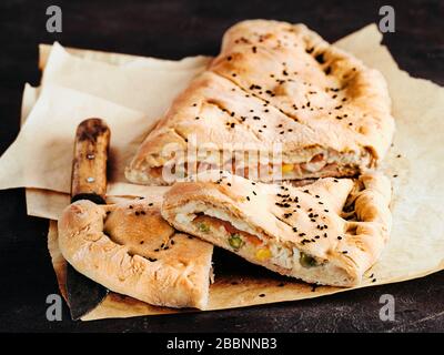 Parfait calzone de pizza fermée maison sur des feuilles de papier de cuisson sur fond noir. Couper de savoureuses pizzas italiennes fermées avec du fromage, de la viande, des légumes, du sésame noir habillé. Espace de copie pour le texte Banque D'Images