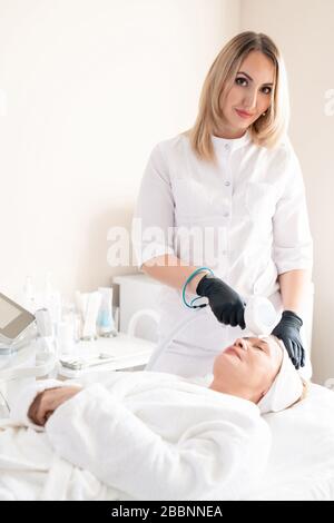 Portrait du contenu jeune cosmologiste dans des gants chirurgicaux utilisant un appareil de beauté pour nettoyer la peau de la femme mûre Banque D'Images