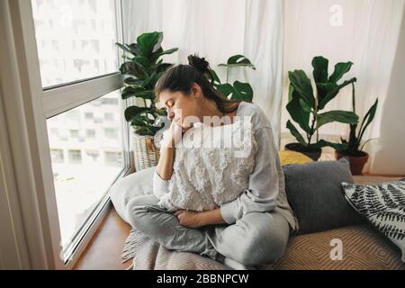 Triste fille hipster tenant l'oreiller, assis à la maison pendant la quarantaine. Isolement à domicile pour prévenir l'épidémie de virus. Jeune femme dans la chambre moderne avec plante Banque D'Images