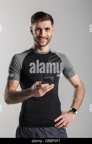 Image d'un sportif dérasé à l'aide d'un téléphone portable et souriant isolé sur fond gris Banque D'Images