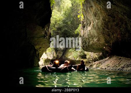Trois personnes portant des phares et flottant sur les anneaux gonflables approchent de l'entrée d'une grotte. Banque D'Images
