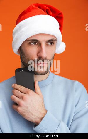 Image d'un jeune homme sérieux à noël chapeau de santa posé isolé sur fond de mur orange tenant le téléphone mobile. Banque D'Images