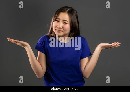 Jeune fille asiatique incertaine dans le hoodie blanc shpettes ses bras, fait le geste de je ne sais pas, soins, ne peut rien aider. Fond gris studio Banque D'Images