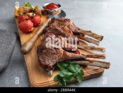 Côtelettes d'agneau rôties sur une planche en bois avec pommes de terre à sauce rouge et tomates Banque D'Images