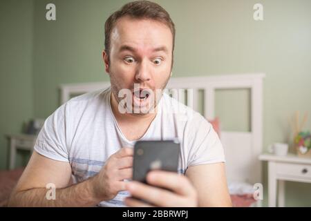 Jeune homme excité avec de grands yeux regardant à son téléphone. L'étonnement s'est fait sentir lors de la lecture de quelque chose sur le smartphone. Banque D'Images