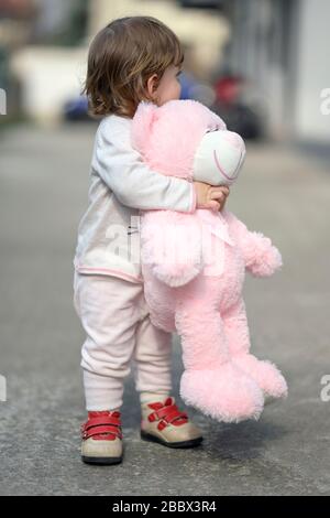 Une jeune fille portant un pantalon et un chemisier roses et des chaussures rouges embrasse un ours en peluche tout en cachant son visage Banque D'Images