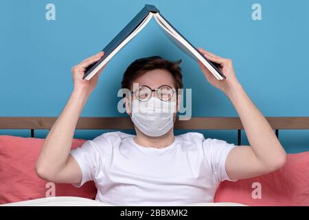 Jeune homme caucasien dans des lunettes et un masque médical lisant un roman dans sa chambre étant seul en quarantaine ou ayant une grippe. Studio tourné Banque D'Images