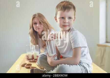 Petit garçon souriant caucasien assis sur la table de la cuisine et manger des biscuits au gruau avec du lait. Petit déjeuner sain avant l'école et la maternelle à la maison. Banque D'Images