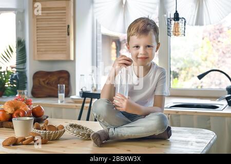 Petit garçon souriant caucasien assis sur la table de la cuisine et manger des biscuits au gruau avec du lait. Petit déjeuner sain avant l'école et la maternelle à la maison. Banque D'Images
