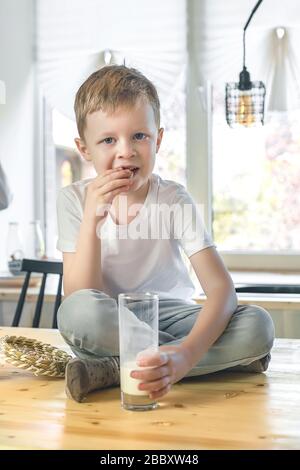 Petit garçon souriant caucasien assis sur la table de la cuisine et manger des biscuits au gruau avec du lait. Petit déjeuner sain avant l'école et la maternelle à la maison. Banque D'Images