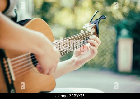 Près du cou de la guitare avec capo dans le parc. Jeune homme assis sur le banc dans le parc jouant de la guitare. Jeune homme séduisant aime la musique live dans Banque D'Images