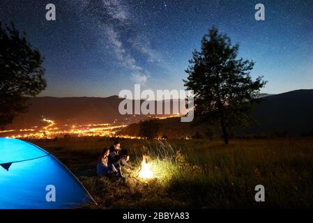 Heureux couple randonneurs se reposant au feu de camp près de la tente touristique bleue éclairée sous le ciel de nuit incroyable plein d'étoiles et voie lactée. Sur l'arrière-plan ciel étoilé, montagnes, grand arbre et ville lumineuse Banque D'Images