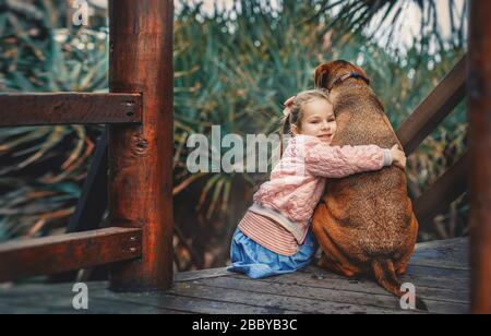 Une jolie petite fille hople un grand chien hongrois vizsla assis ensemble sur les escaliers en bois. Enfant et animal de compagnie. Banque D'Images