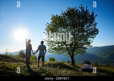 Vue arrière de jeunes touristes de couple se reposant près du camping d'été et grand arbre dans les montagnes au soleil matin. Homme romantique et femme randonneurs tenant les mains. Banque D'Images