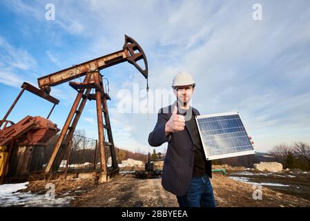 Portrait d'homme d'affaires portant un manteau de sport, un Jean et un casque blanc, tenant un mini module solaire, montrant le pouce vers le haut, debout dans un champ d'huile à côté du cric de pompe à huile. Concept de sources d'énergie alternatives. Banque D'Images