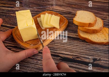 Quelqu'un coupe un peu de beurre avec un couteau d'un morceau dans un plat de beurre en bois, des tranches de fromage et des toasts de blé frais sur un fond en bois. Fermer u Banque D'Images