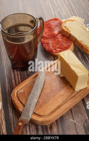 Pain grillé au blé frais avec beurre et saucisse, un plat à beurre en bois avec un morceau de beurre, un couteau avec une poignée en bois et un mug à café en verre sur une boode Banque D'Images