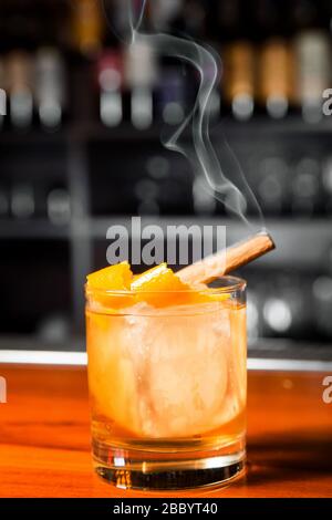 Cocktail orange avec cube de glace et zeste d'orange. Dans le verre est un bâton de cannelle, d'où il y a de la fumée. Banque D'Images