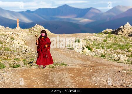 Femme pèlerine dans une robe rouge marchant sur la Kora. Elle fait tourner une roue de prière de la main dans sa main droite. Dans les montagnes arrière du plateau tibétain. Banque D'Images