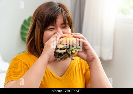 Une jeune femme asiatique affamée en surpoids qui tient du hamburger, qui a faim à tout moment et qui mange trop, qui mange de la gluttonie et des dindes. Son style de vie est de manger de la nourriture rapide A Banque D'Images