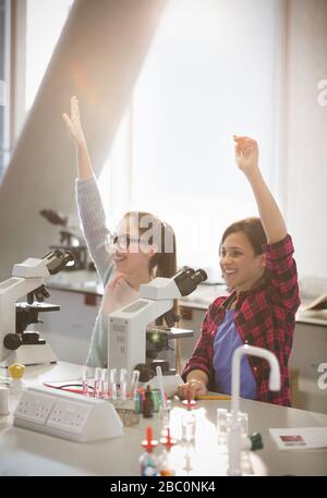 Des jeunes filles avides élèvent les bras derrière des microscopes en salle de classe de laboratoire Banque D'Images