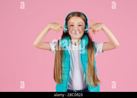 Une petite fille avec casque explose la gomme à mâcher rose sur un fond rose. Banque D'Images