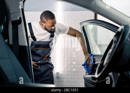 Jeune employé africain du centre de soins de la voiture en salopette et t-shirt tenant générateur de vapeur et tissu en microfibre pour le nettoyage du salon de la voiture. Voiture Banque D'Images