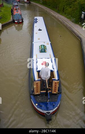 Vue depuis un pont routier en haut d'un bateau à narrowboat qui passe sous le canal Grand Union avec un homme au timon. Banque D'Images