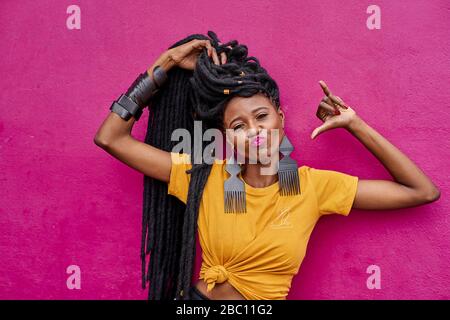 Portrait de femme avec de longs readlocks faisant signe shaka devant un mur rose Banque D'Images