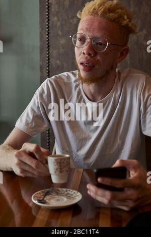 Albino homme avec des verres ronds ayant un café et de vérifier son téléphone dans une cafétéria Banque D'Images