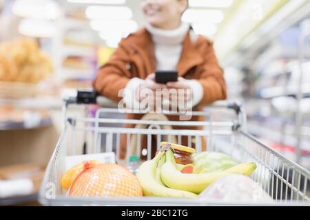 Femme avec smartphone poussant panier dans le supermarché Banque D'Images